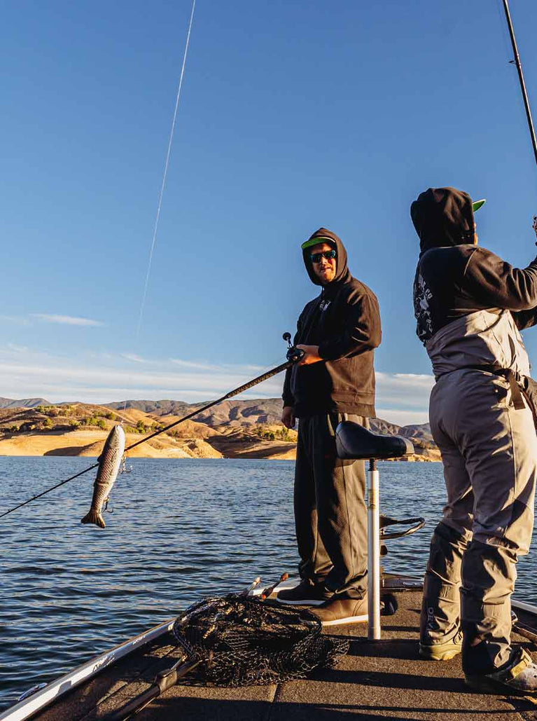 Two men on a boat with one holding a fish, against a clear blue sky.