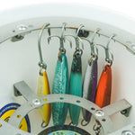 A close-up of a white circular fishing lure storage ring holding six metal treble hooks with colorful fishing lures in yellow, teal, green, purple, silver, and orange. A spool of line is partially visible at the bottom.
