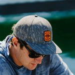 Man wearing a camouflage cap with a logo and sunglasses, sitting by water.