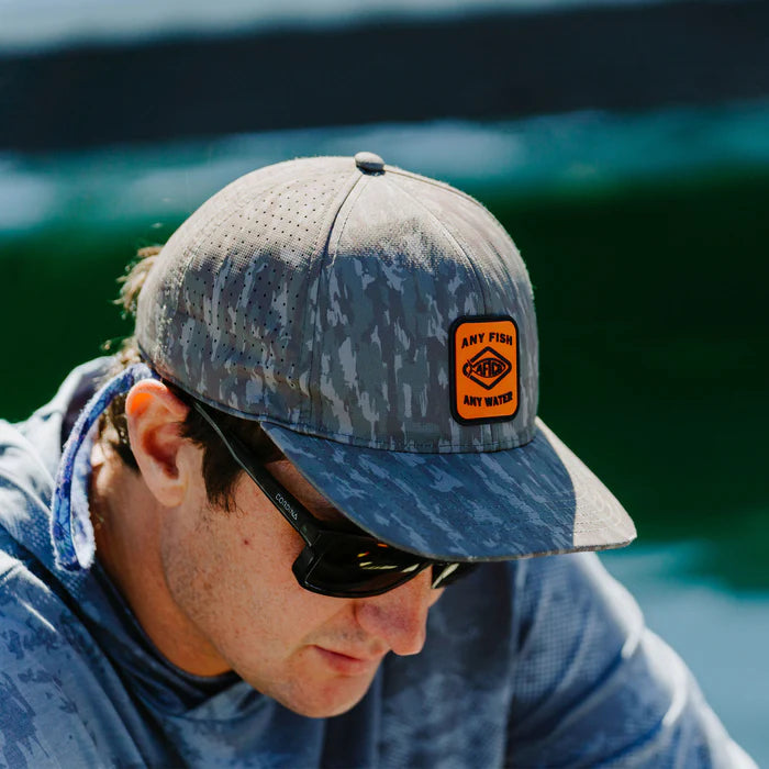 Man wearing a camouflage cap with a logo and sunglasses, sitting by water.