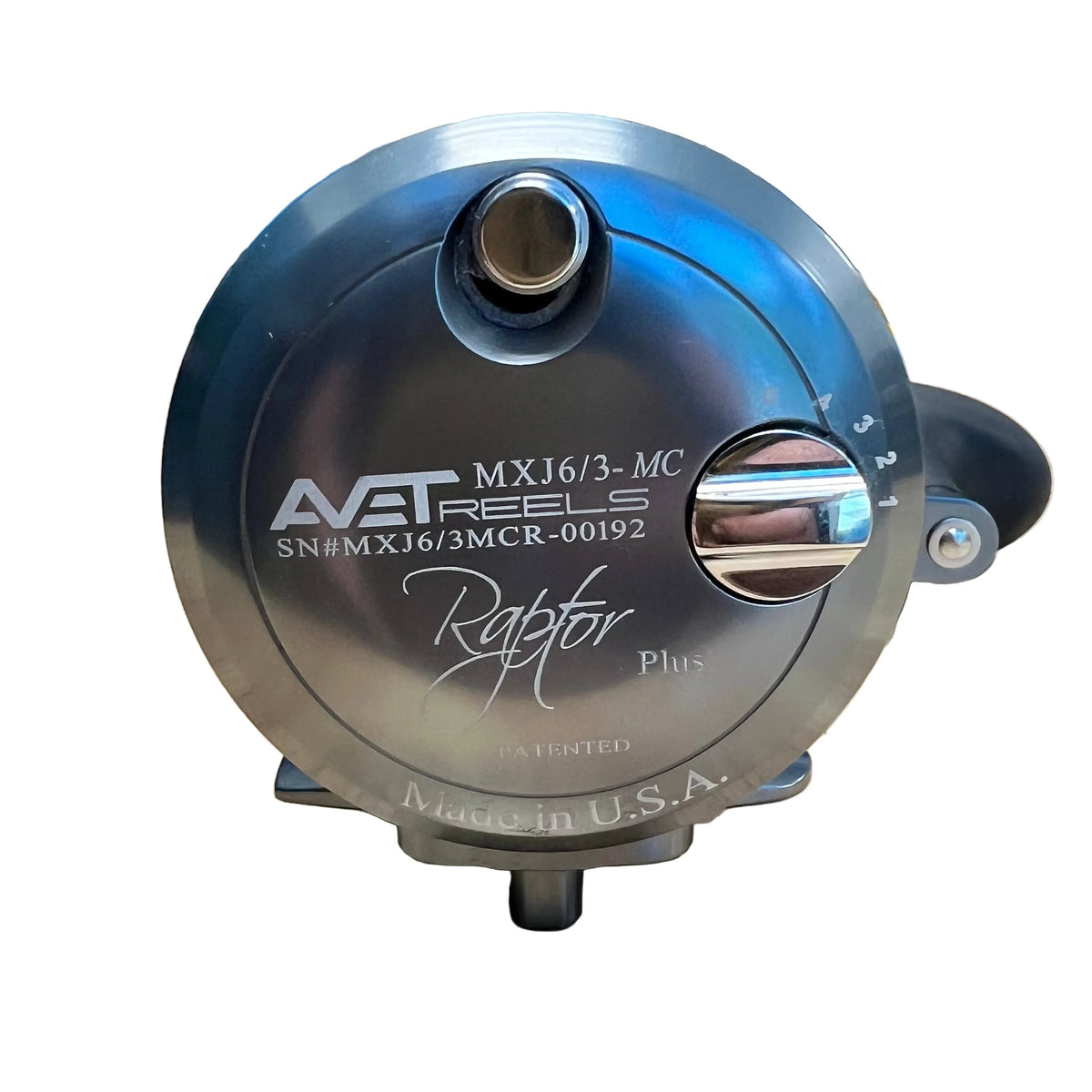 A metallic silver Avt Reels Raptor Plus fishing reel sitting on a light-colored wooden tabletop, with a workshop or garage background.