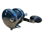 A close-up of a black and dark gray baitcasting fishing reel with a black handle, resting on a light-colored wooden table. The reel features silver metal knobs and the brand name 'AGAPE' is visible on the side.